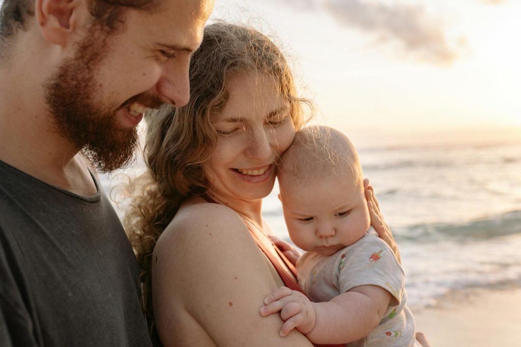 A joyful family moment with parents holding their baby by the ocean at sunset.